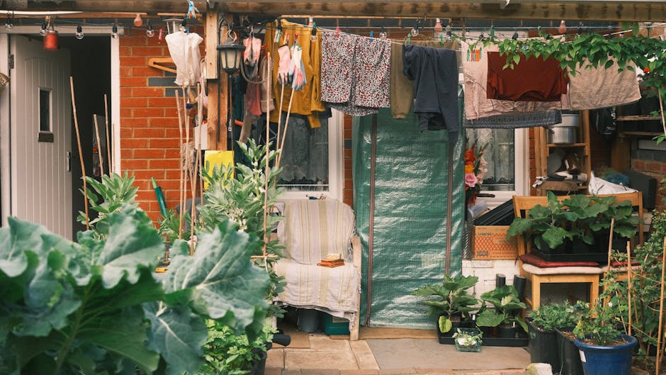 A small, functional backyard garden featuring various gardening tools and potted plants arranged along a brick wall. In the foreground, large green leaves of vegetable plants, possibly squash or courg